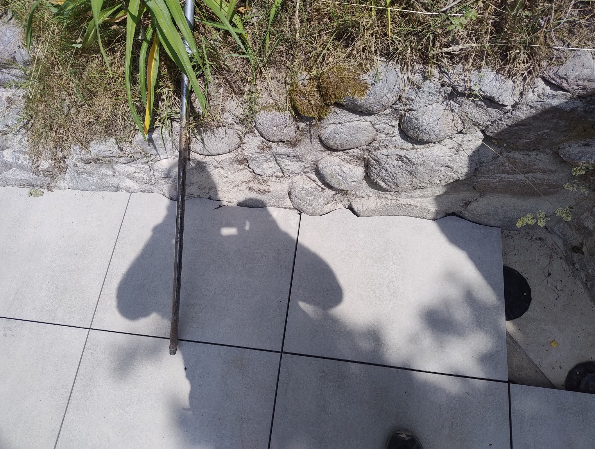 Jasnoszare płytki ułożone na zewnątrz, przy kamiennym murku z roślinnością. Widoczny cień osoby i narzędzia. Płytki ułożone równo, z ciemnymi fugami.