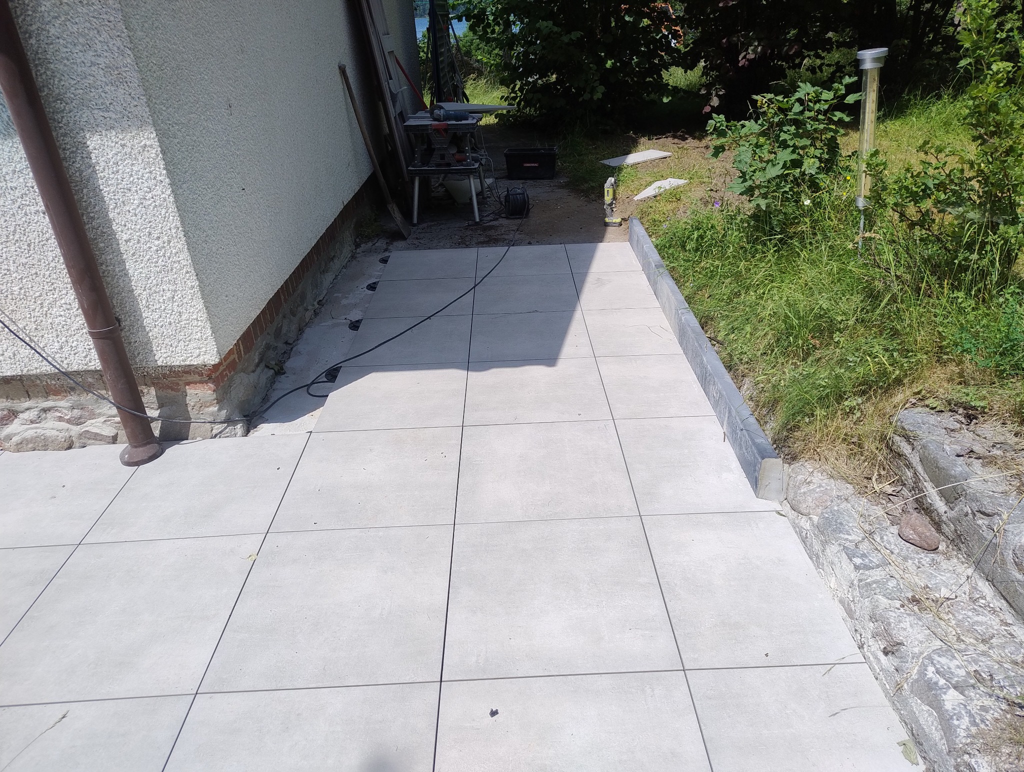 Ułożona, jasnoszara, kwadratowa terakota na zewnątrz budynku. W tle widoczna piła do cięcia płytek i narzędzia. Częściowo ukończona realizacja.