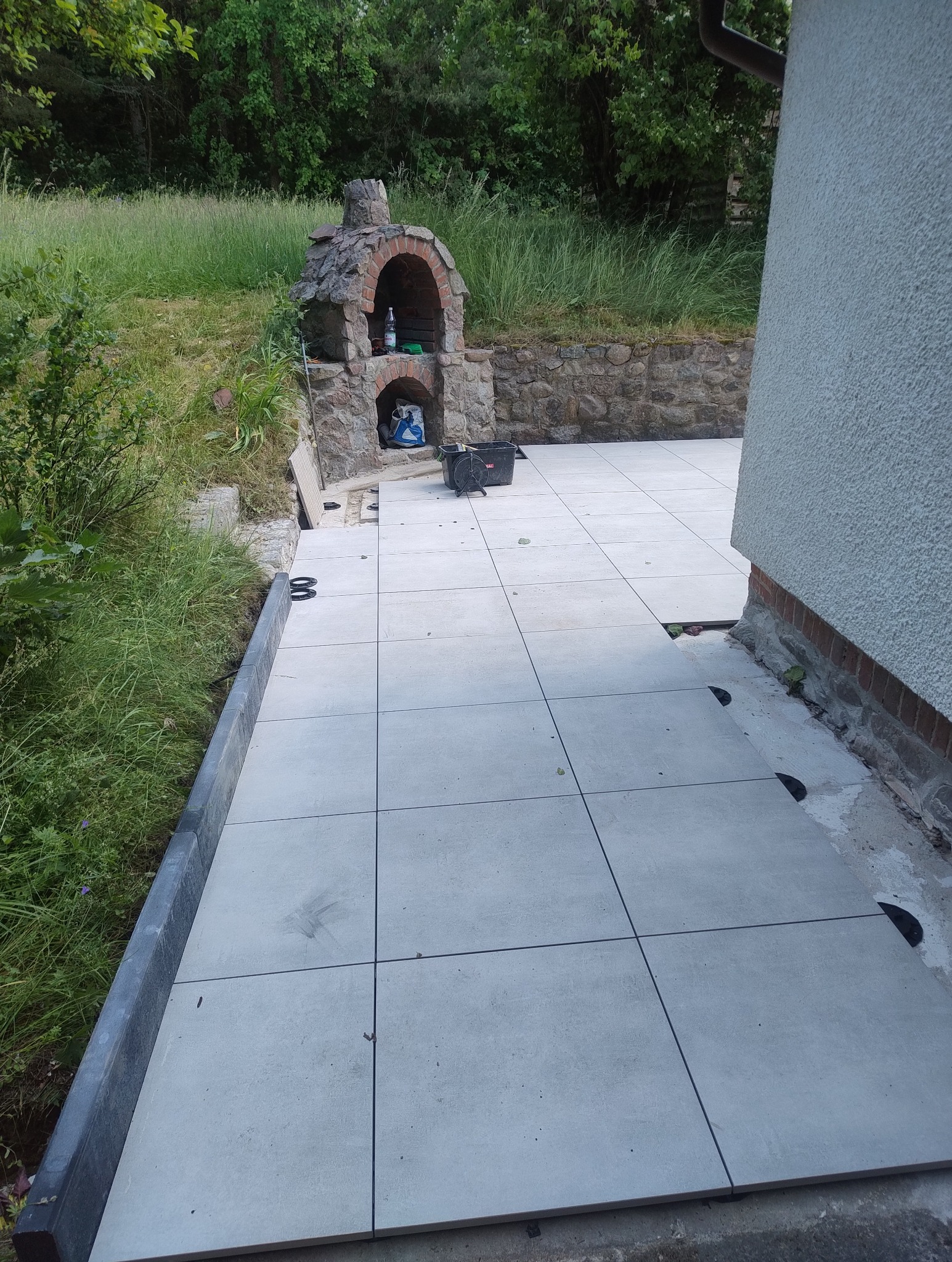 Układanie jasnoszarej terakoty na zewnątrz, widoczny fragment budynku i murowany grill. Częściowo ułożona podłoga z terakoty na wspornikach.