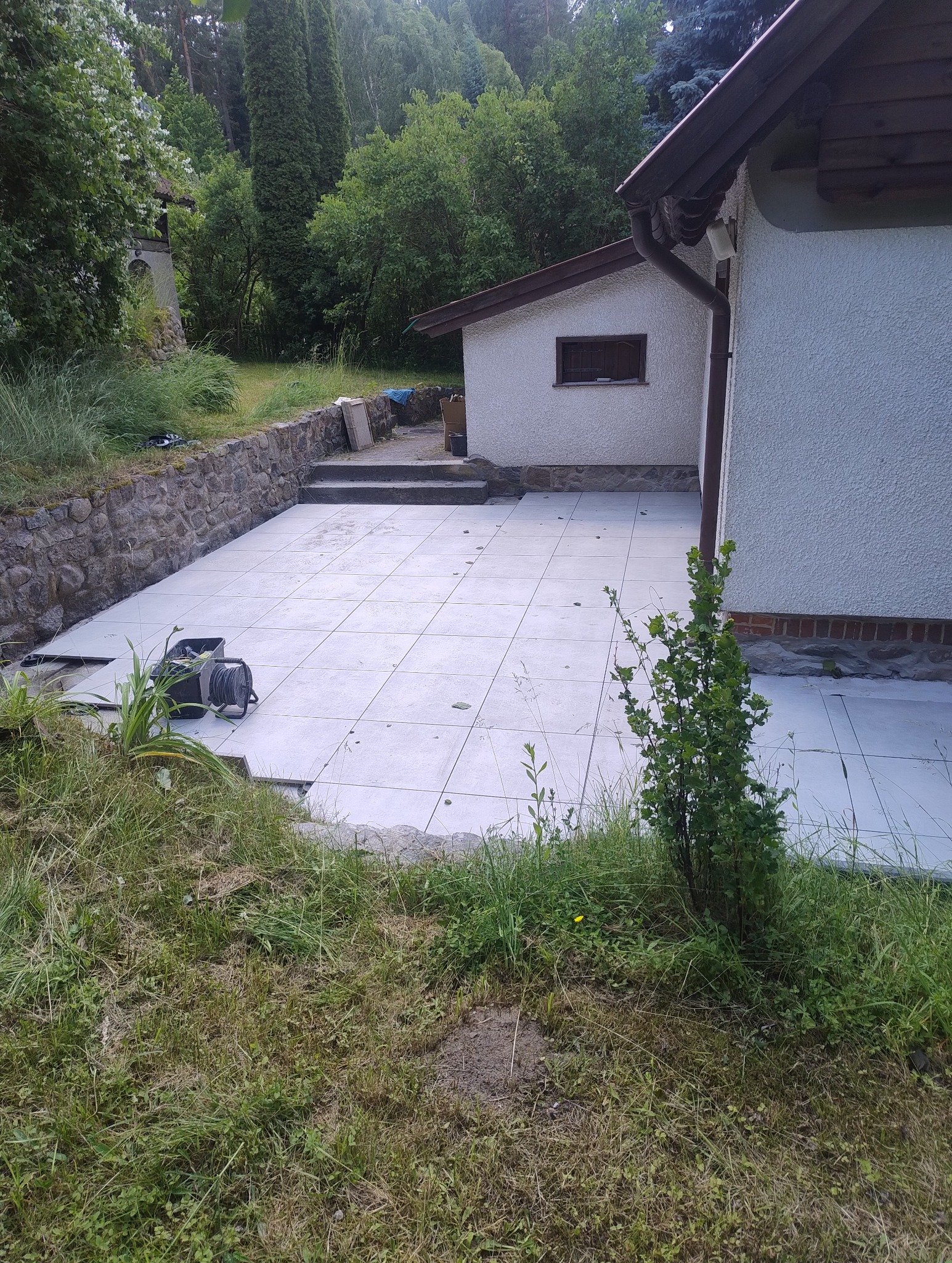 Jasnoszara, kwadratowa płytka ułożona na zewnątrz budynku, tworząca taras. Widoczne narzędzia i trawa. Częściowo ukończona realizacja.