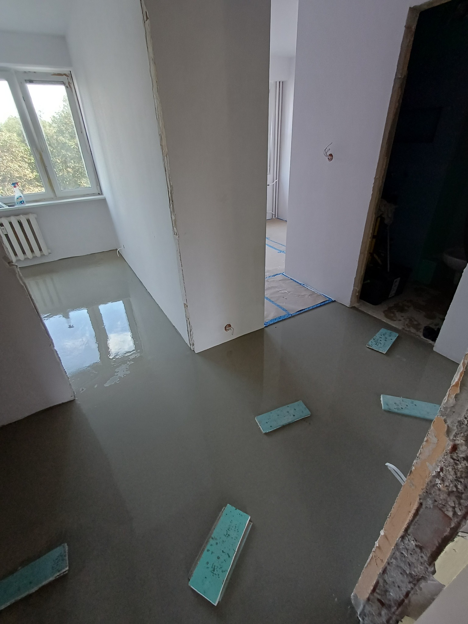 Wnętrze w trakcie wylewania posadzki samopoziomującej, widoczne fragmenty ścian i okno z widokiem na zieleń. Na podłodze leżą gąbki. Widoczny stan surowy.