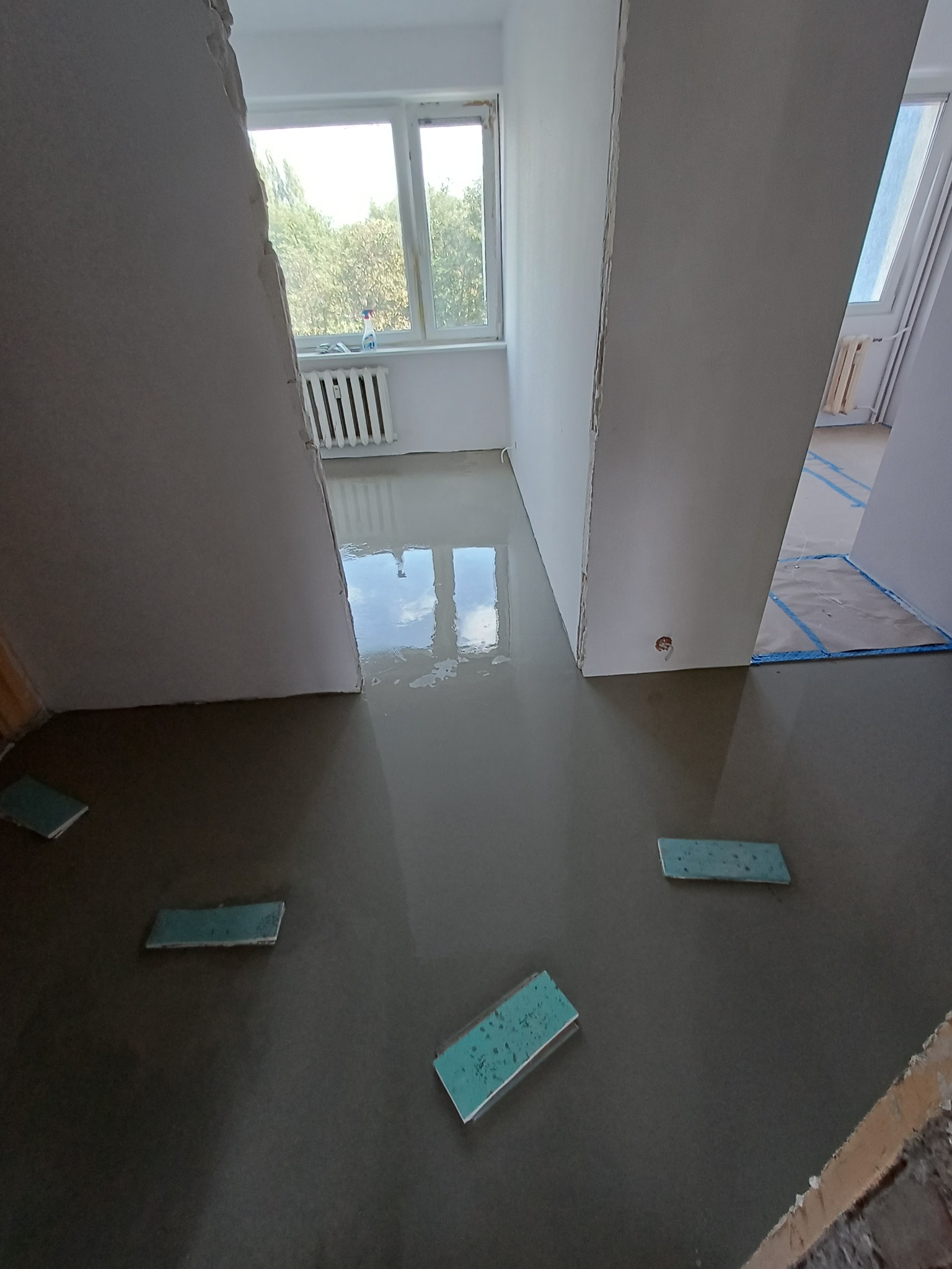 Wylewka samopoziomująca w trakcie realizacji. Widoczne fragmenty ścian, okno z widokiem na zieleń i poziomice. Świeża, mokra powierzchnia wylewki odbija światło.
