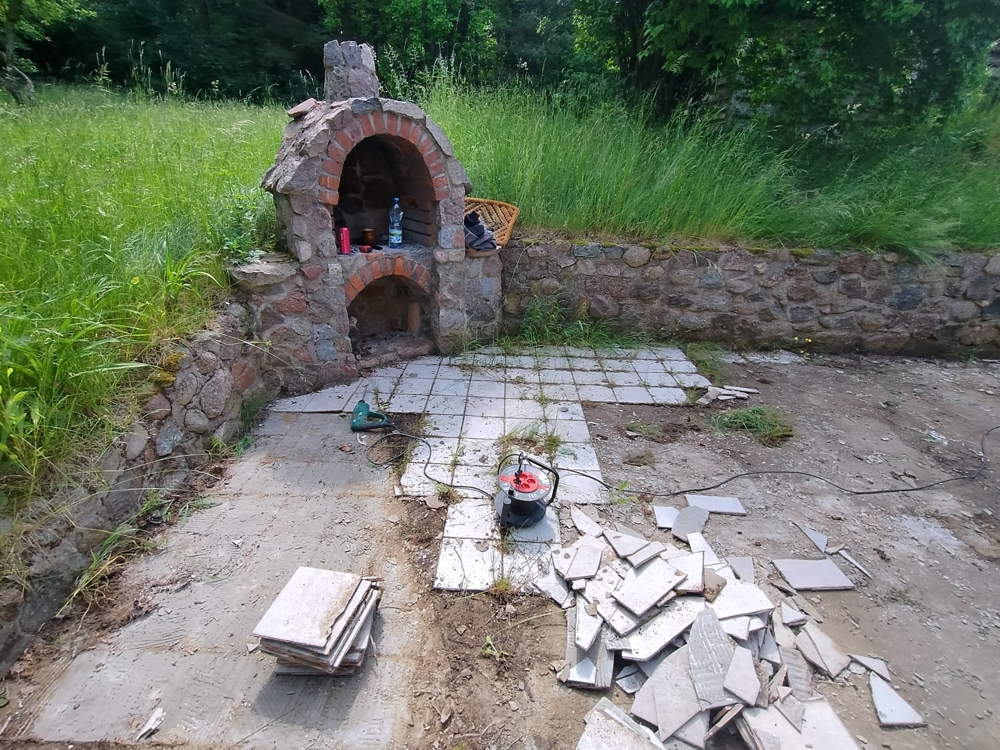 Rozbiórka płytek na zewnątrz przy kamiennym grillu. Widoczne narzędzia i sterta potłuczonych płytek. Częściowo usunięte płytki odsłaniają podłoże. Prace w toku.