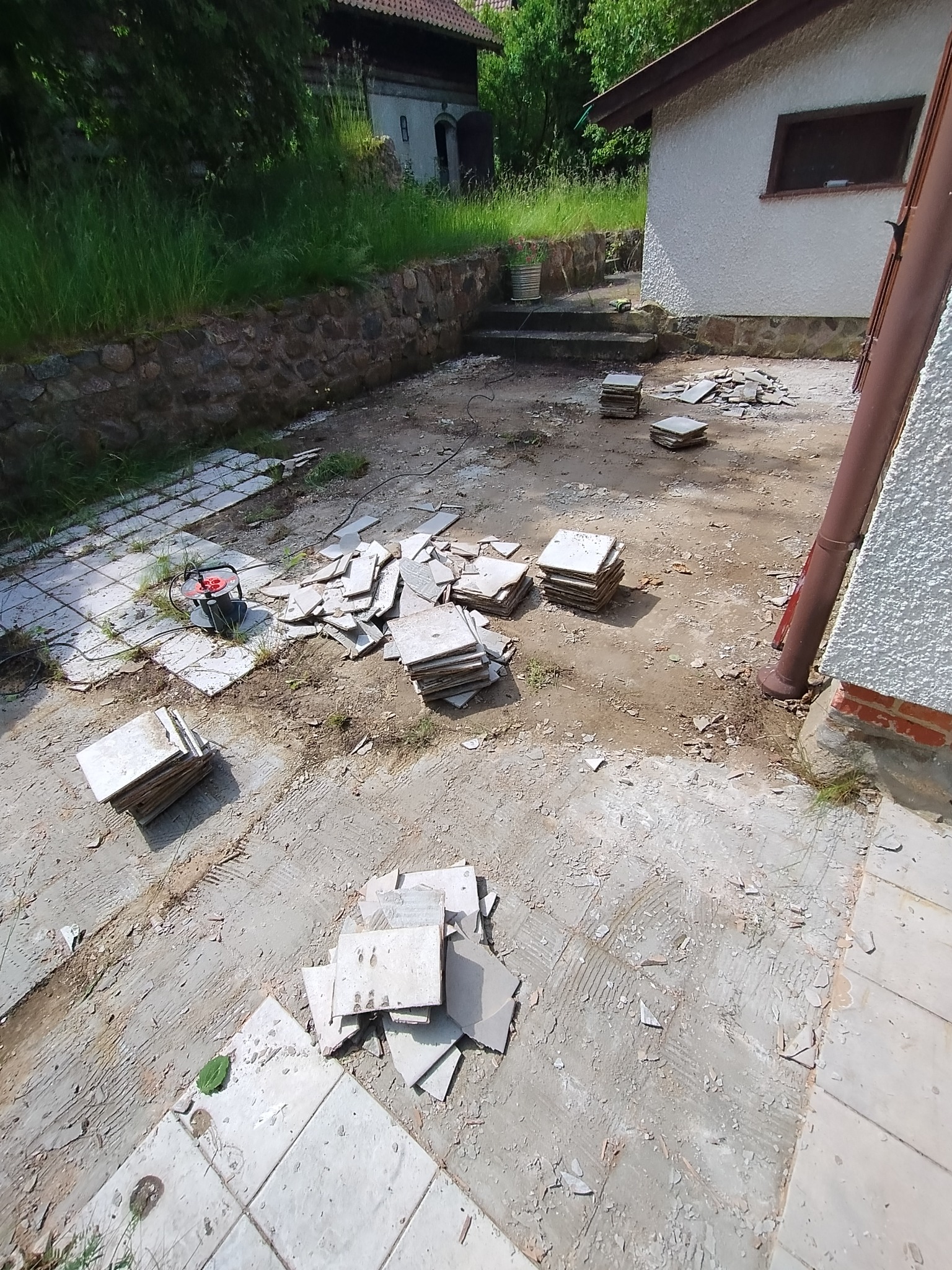 Demontaż starej terakoty na zewnątrz budynku. Widoczne narzędzie do cięcia płytek, stosy zdjętych płytek i fragmenty starej zaprawy. Prace przygotowawcze do położenia nowej nawierzchni.