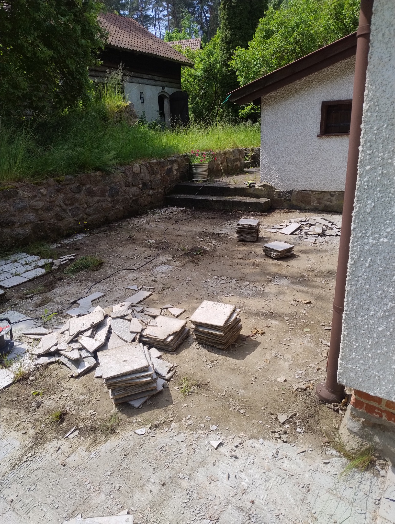 Przygotowanie do ułożenia płytek na zewnątrz budynku. Stosy płytek i gruz na ziemi, widoczne schody i budynek w tle. Prace glazurnicze w toku.