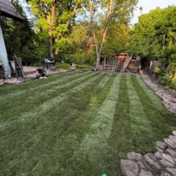 Układanie trawy z rolki w ogrodzie z kamienną ścieżką. Dwóch pracowników montuje pasy, tworząc wzór w paski. W tle widoczne drzewa i budynek.
