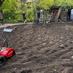 ZAKAQUA Michał Zakrzewski - Czerwony glebogryzarka Cedrus GLX480 na przygotowanym podłożu w ogrodzie, widoczni pracownicy w tle, narzędzia i drewno składowane obok.