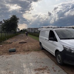 ZAKAQUA Michał Zakrzewski - Etap przygotowania terenu pod trawnik: rozłożona instalacja nawadniająca, narzędzia i biały samochód dostawczy na tle pochmurnego nieba i zabudowy miejskiej.