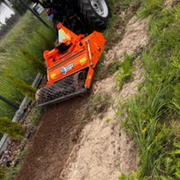 Traktor z glebogryzarką wyrównuje ziemię na działce, przygotowując ją pod przyszły trawnik. Widoczne świeżo przekopane pasy ziemi i fragment ogrodzenia.