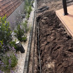 ZAKAQUA Michał Zakrzewski - Instalacja nawadniania w ogrodzie: rura w wykopanym rowie, obok rabata z kamykami, roślinami i lampą. W tle drewniany płot i fragment tarasu.