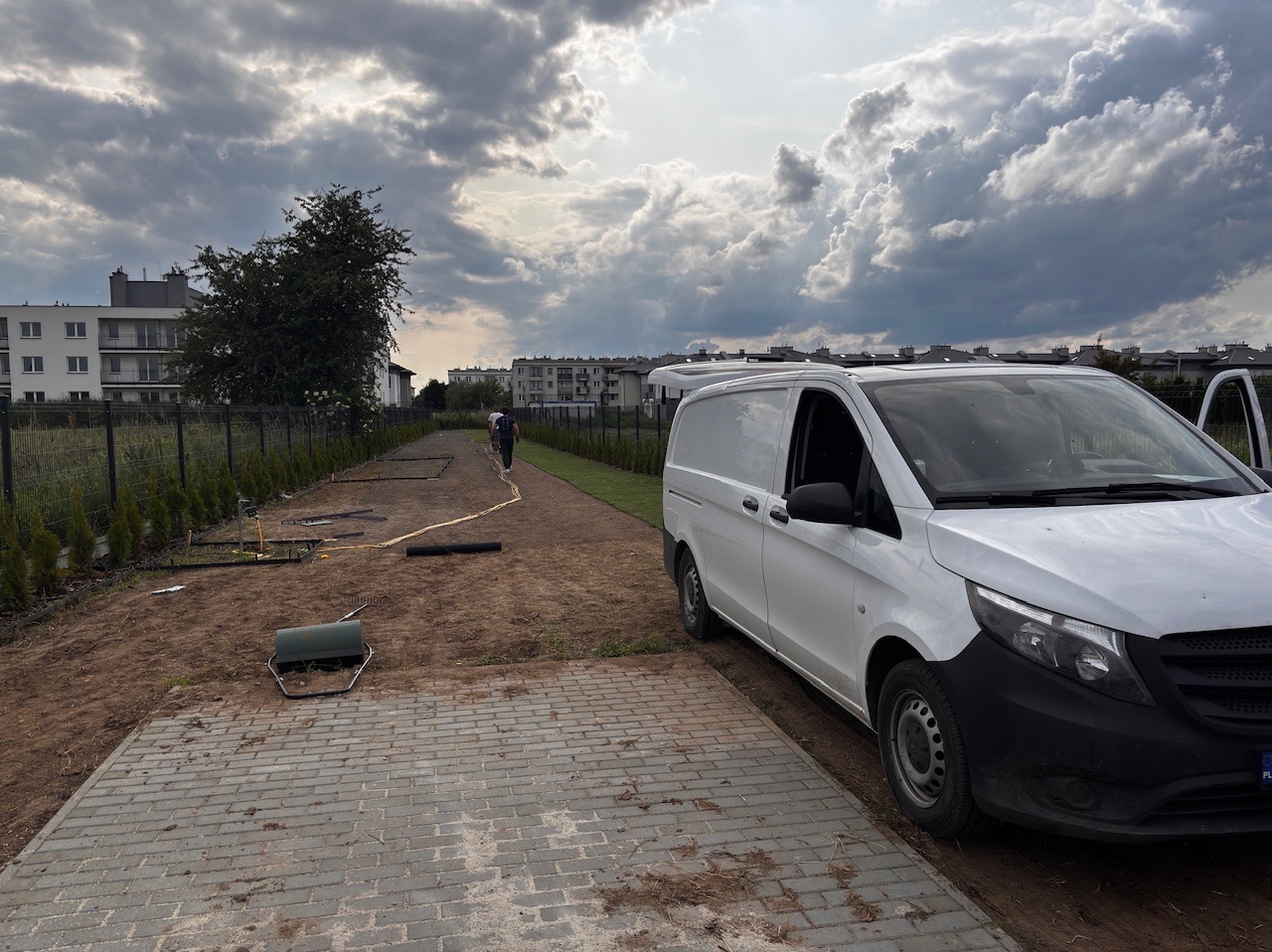 Etap przygotowania terenu pod trawnik: rozłożona instalacja nawadniająca, narzędzia i biały samochód dostawczy na tle pochmurnego nieba i zabudowy miejskiej.