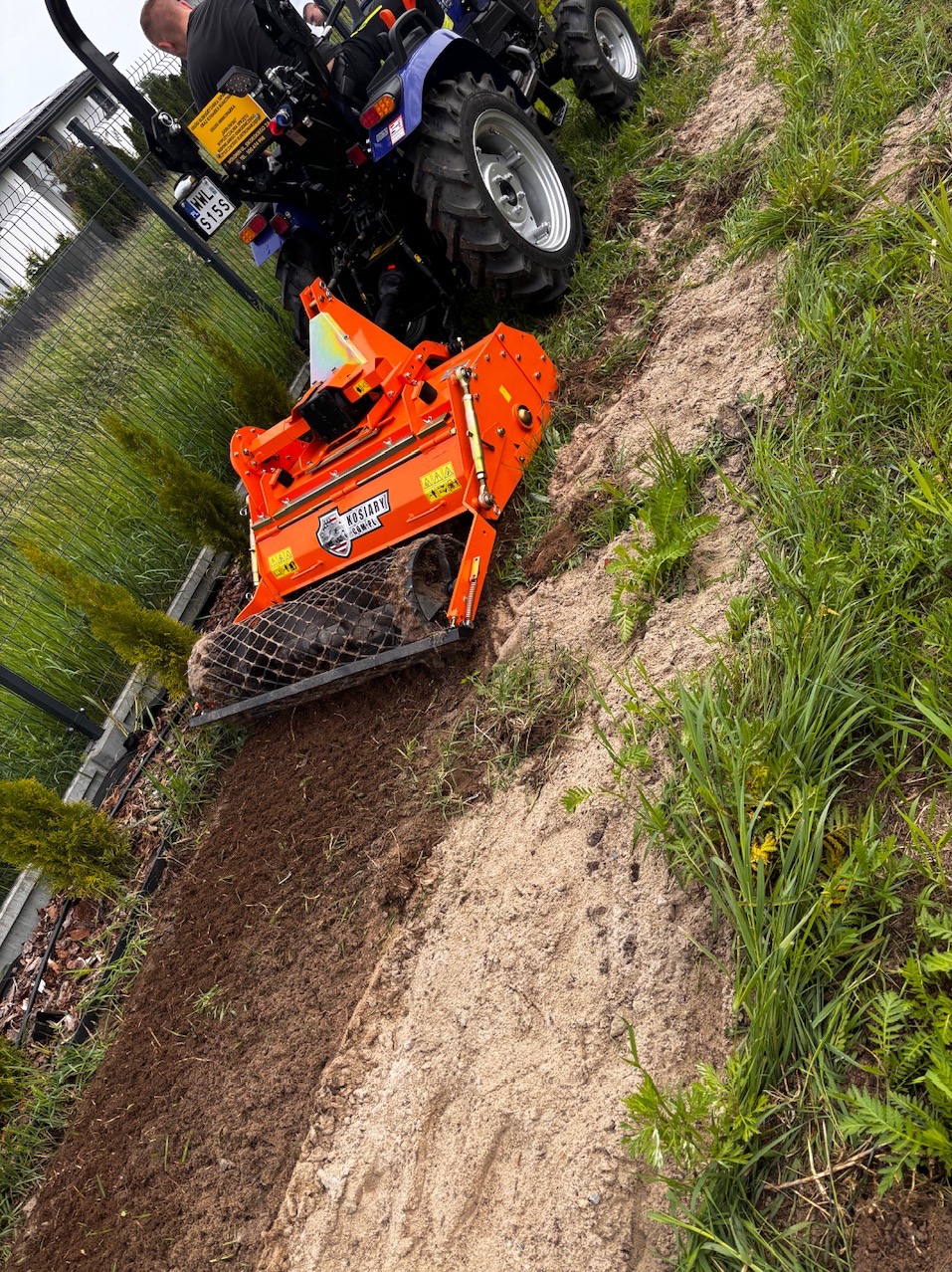 Traktor z glebogryzarką wyrównuje ziemię na działce, przygotowując ją pod przyszły trawnik. Widoczne świeżo przekopane pasy ziemi i fragment ogrodzenia.