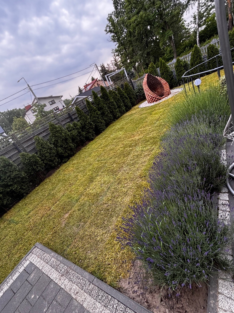 Ujęcie na ogród z trawnikiem, rzędem tuj i dekoracyjną konstrukcją z cegieł. Fioletowa lawenda wzdłuż ścieżki dodaje koloru. Pochmurne niebo w tle.