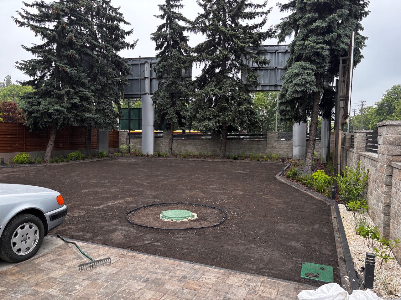 Przygotowany teren pod trawnik z widocznymi drzewami iglastymi, obrzeżami i studzienką kanalizacyjną. Grabie leżą na kostce brukowej. Częściowo widoczny samochód.