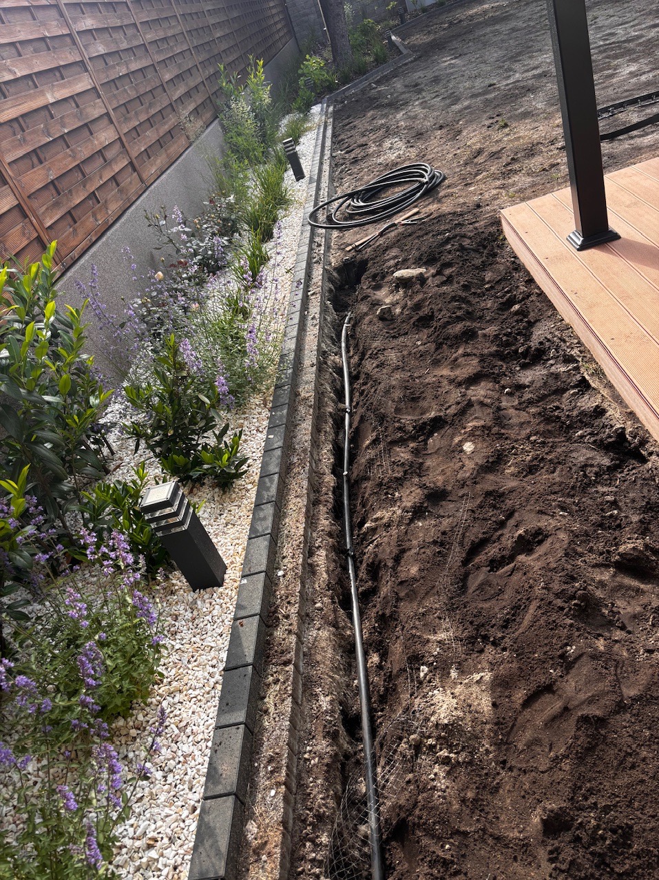 Instalacja nawadniania w ogrodzie: rura w wykopanym rowie, obok rabata z kamykami, roślinami i lampą. W tle drewniany płot i fragment tarasu.