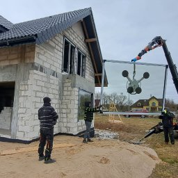 ADBUD - Montaż okna na budowie domu jednorodzinnego z użyciem dźwigu i przyssawki. Widoczni pracownicy w odblaskowych kamizelkach. Dom w stanie surowym.