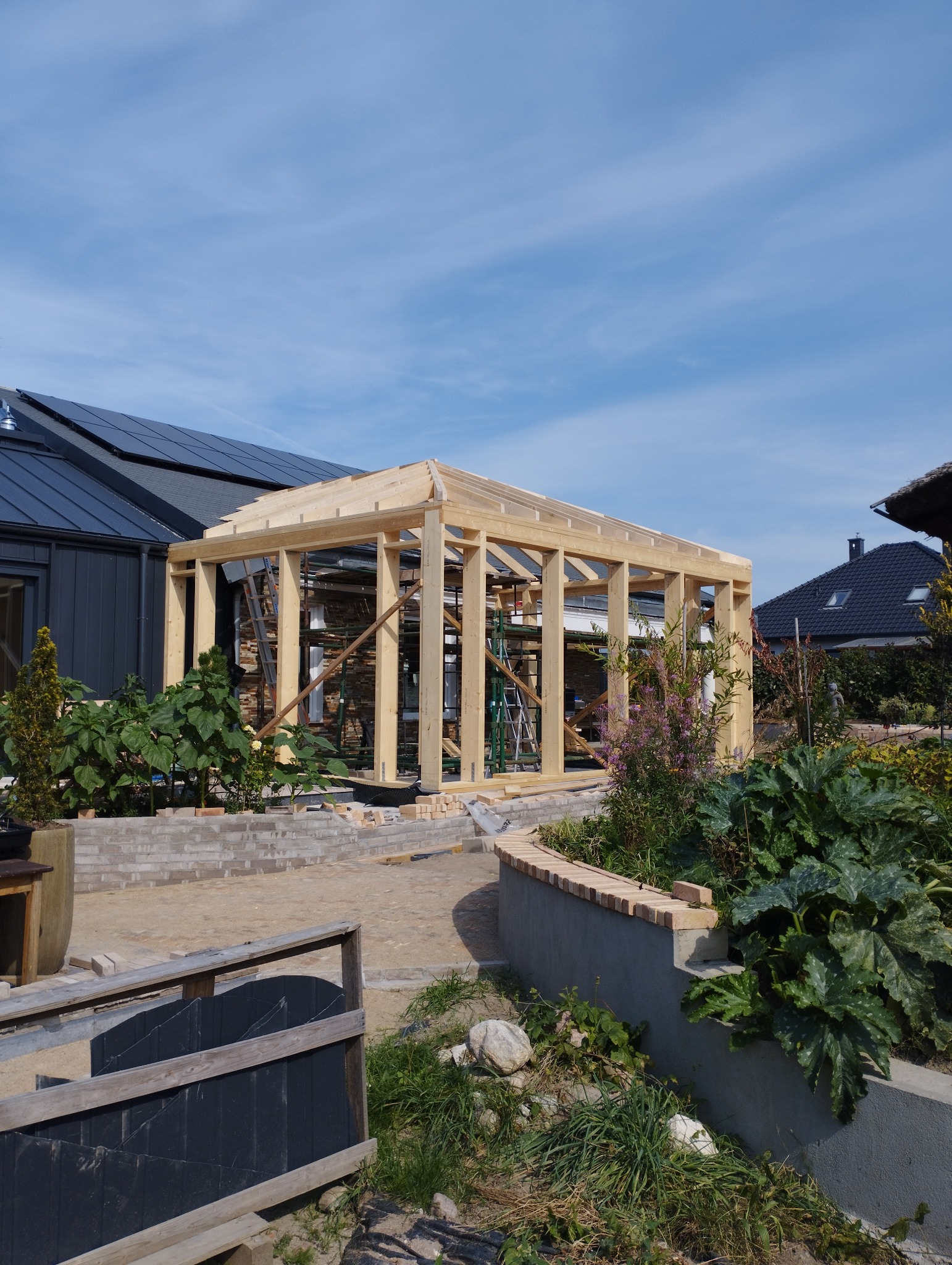 Drewniana konstrukcja altany w trakcie budowy, widoczne rusztowania i narzędzia, w tle dom z panelami słonecznymi i bujna roślinność ogrodowa.