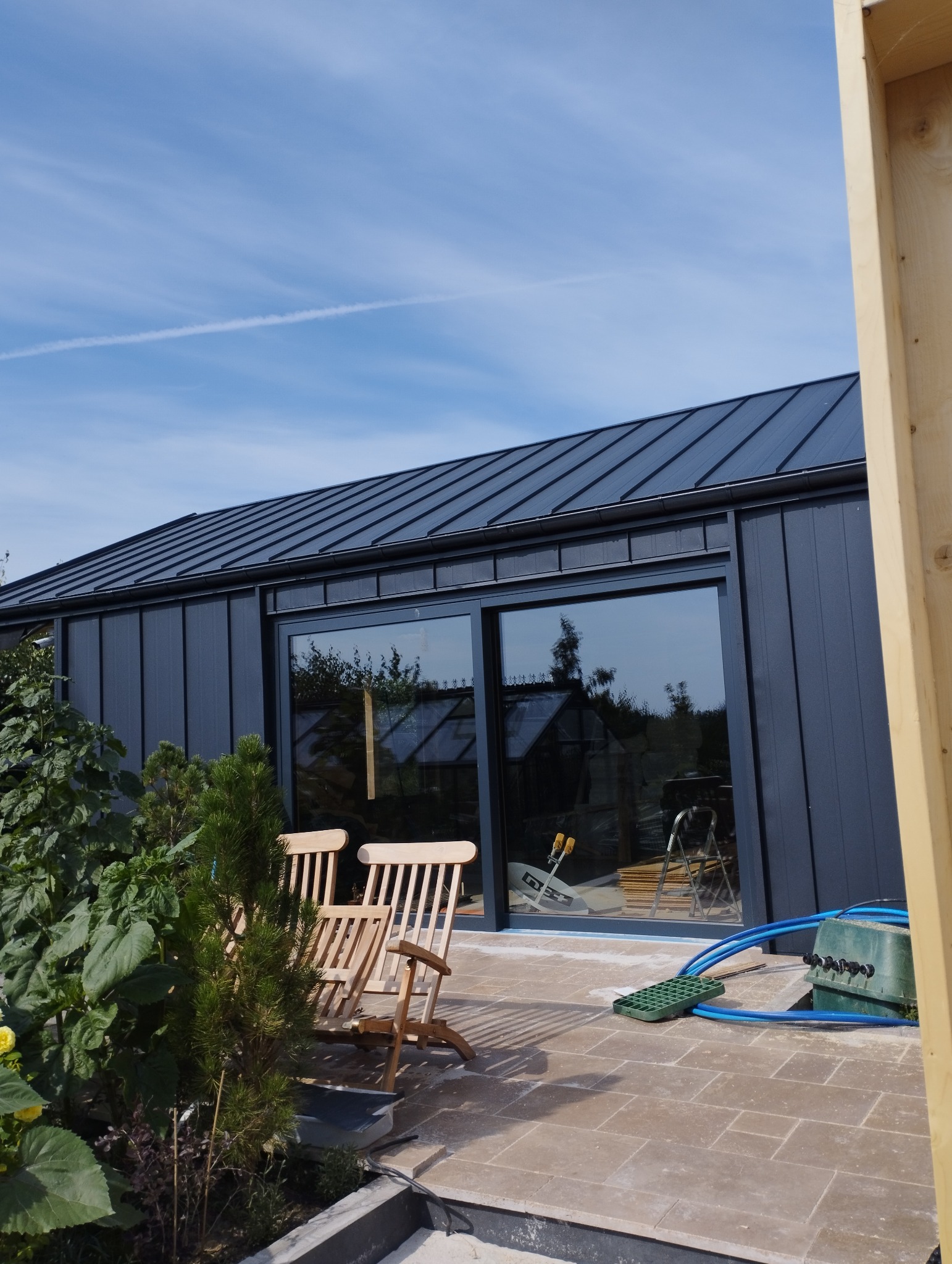 Nowoczesny dom szkieletowy z ciemną blachą na dachu, widoczne drewniane leżaki na tarasie i fragment ogrodu z roślinami.