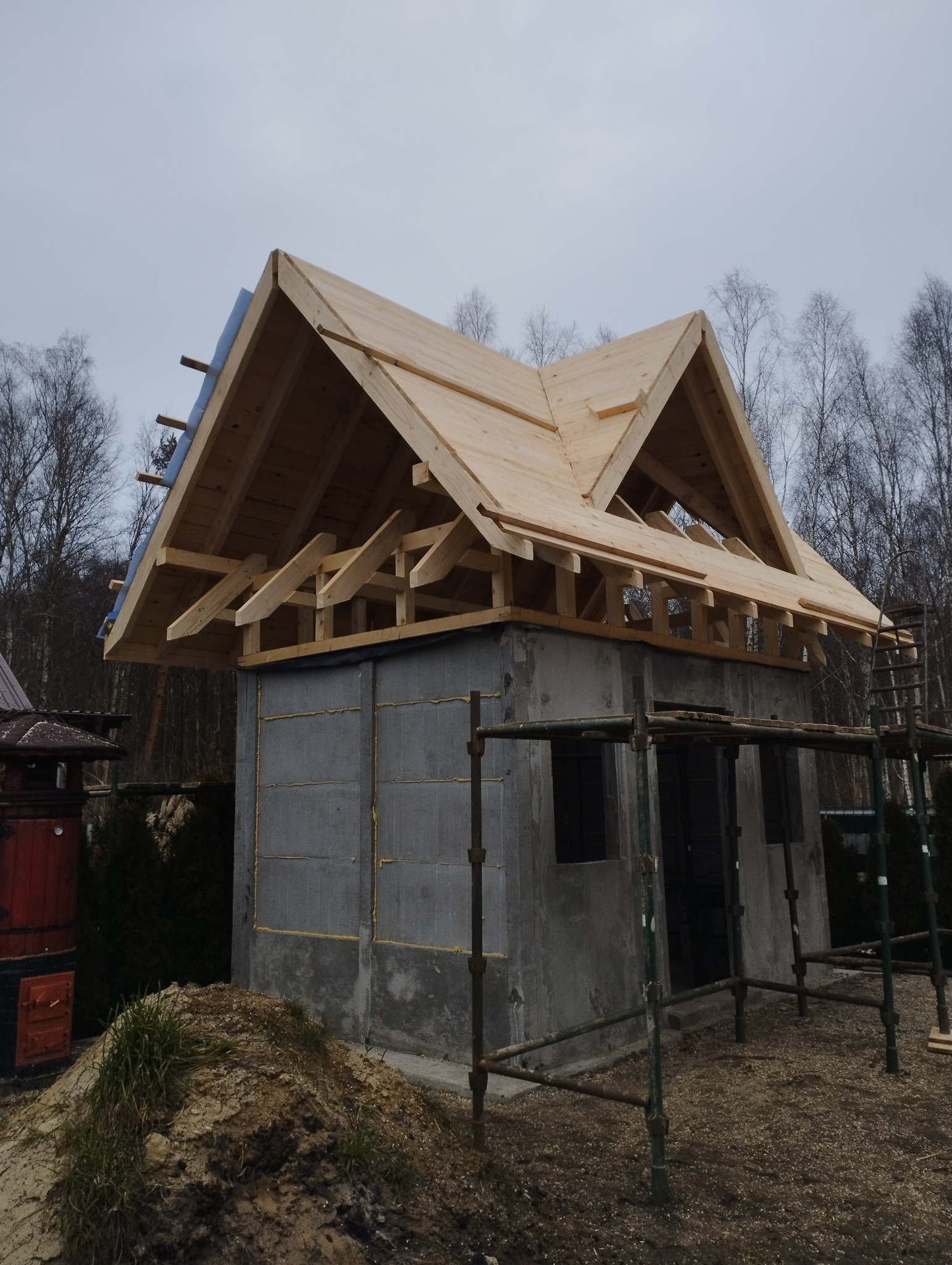 Surowy budynek z betonu komórkowego w trakcie budowy, z widoczną drewnianą konstrukcją dachu i częściowo położonym poszyciem, otoczony rusztowaniem i drzewami bezlistnymi.