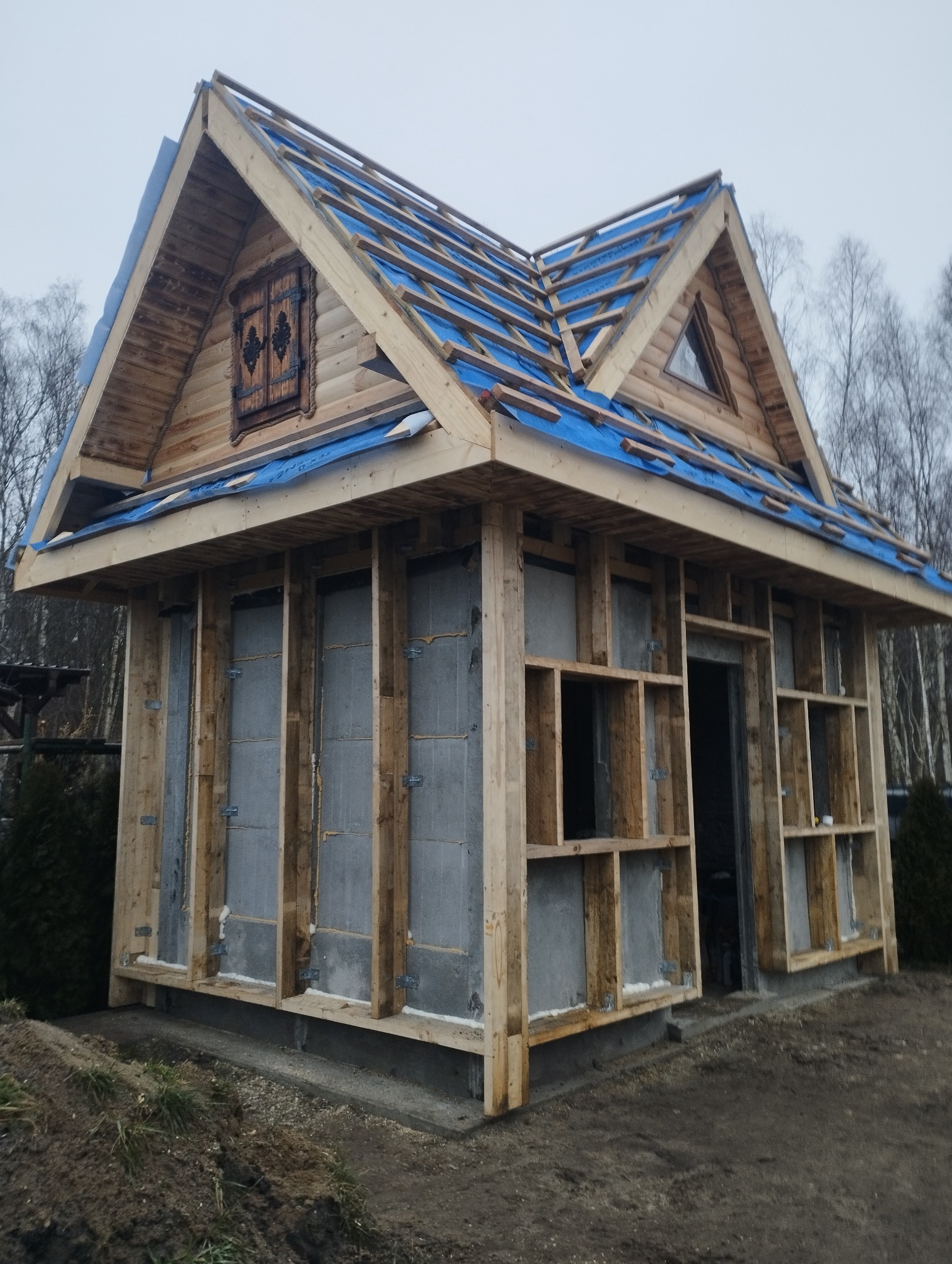 Budowa domu szkieletowego z drewnianą konstrukcją i niebieską membraną dachową, widoczne bloczki betonowe na ścianach, okno z ozdobnymi okiennicami.