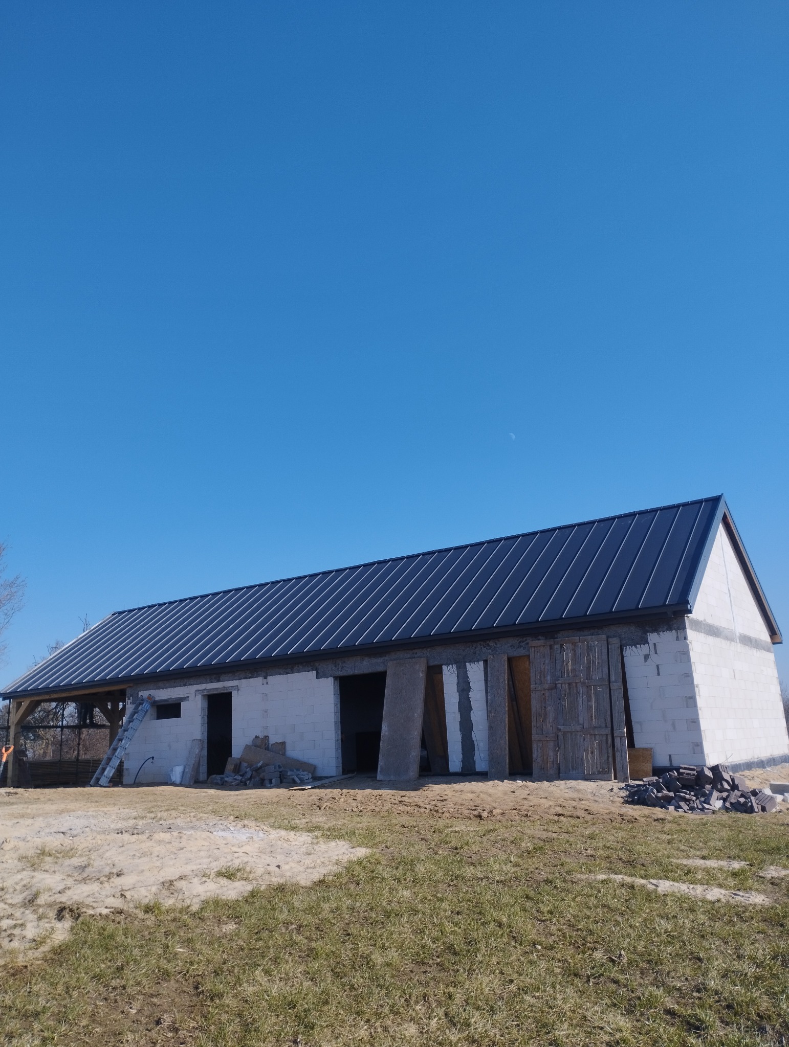 Budynek gospodarczy w trakcie budowy z ciemnym, metalowym pokryciem dachowym, widoczne bloczki betonowe, niezabezpieczone otwory okienne i drzwiowe oraz stos cegieł na zewnątrz.