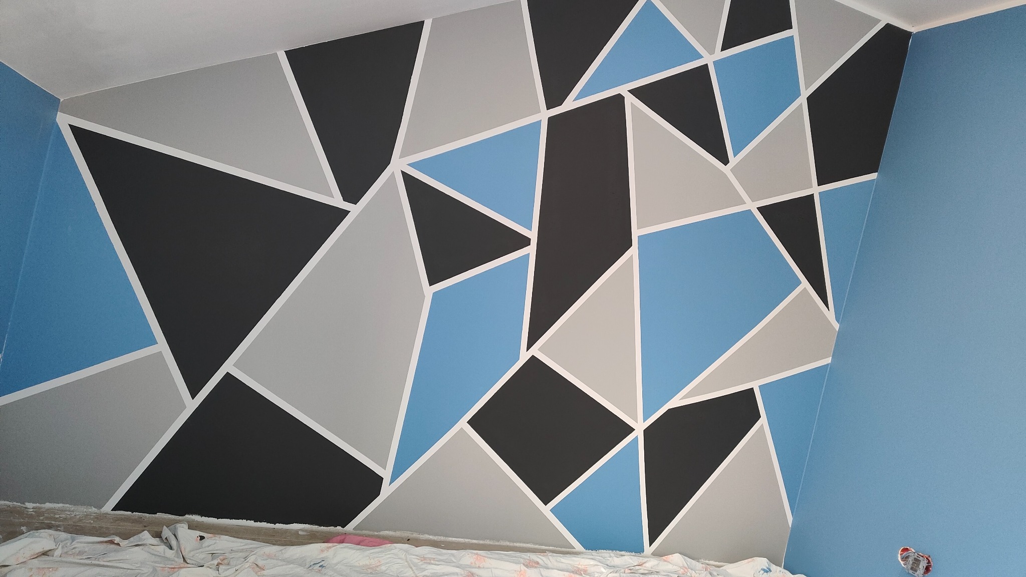 Ściana w pokoju pomalowana w geometryczne wzory z użyciem kolorów: błękitny, szary i czarny, oddzielone białymi liniami. Na podłodze widoczna folia ochronna.