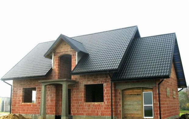 Surowy stan rozbudowy domu jednorodzinnego z czerwonej cegły, z zamontowanym dachem z blachodachówki w kolorze grafitowym, widoczne niezadaszone wejście i otwory okienne.