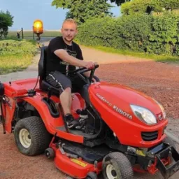 Młody mężczyzna siedzi na czerwonej kosiarce trawnikowej Kubota GR1600II na tle zieleni i ścieżki. Kosiarce towarzyszy żółte światło ostrzegawcze.