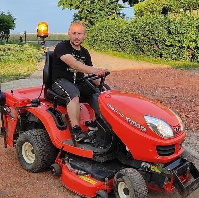 Młody mężczyzna siedzi na czerwonej kosiarce trawnikowej Kubota GR1600II na tle zieleni i ścieżki. Kosiarce towarzyszy żółte światło ostrzegawcze.