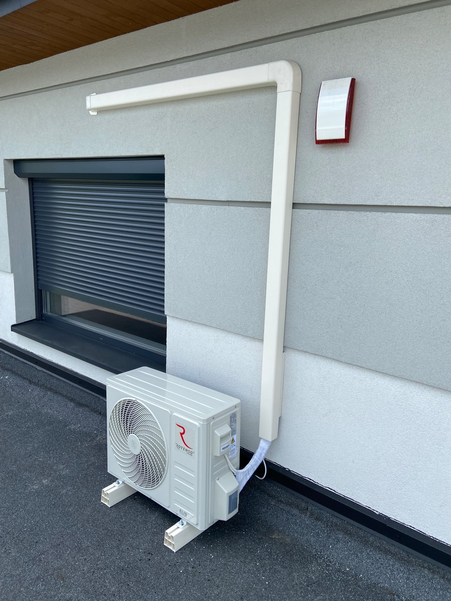 Zewnętrzna jednostka klimatyzatora marki Rotenso, model Elis, zamontowana na wspornikach z osłoną instalacji na elewacji budynku z zamkniętą roletą okienną i czujnikiem.