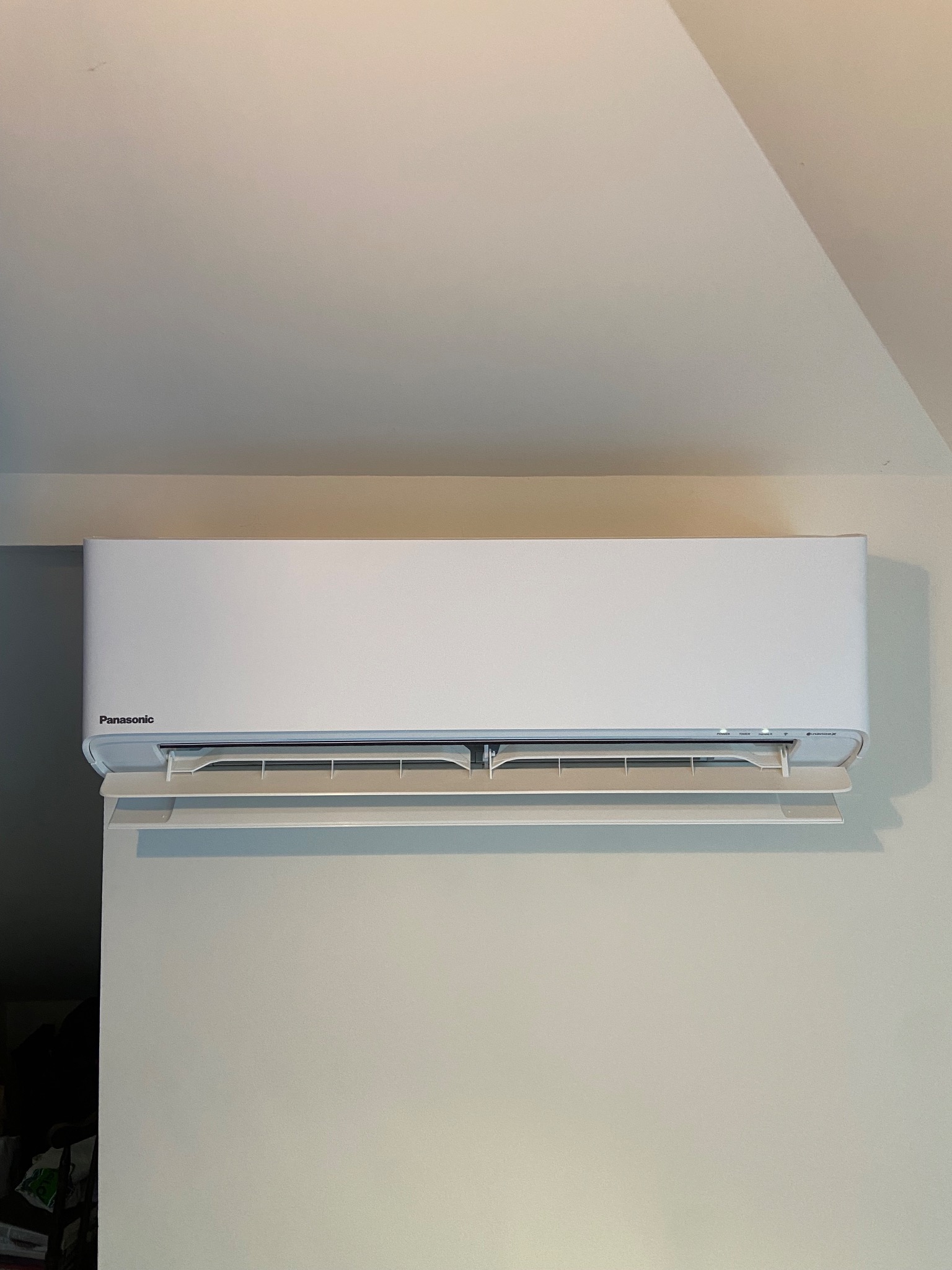 Biały klimatyzator Panasonic zamontowany na jasnej ścianie, widok z dołu, widoczne detale obudowy i regulowane żaluzje.