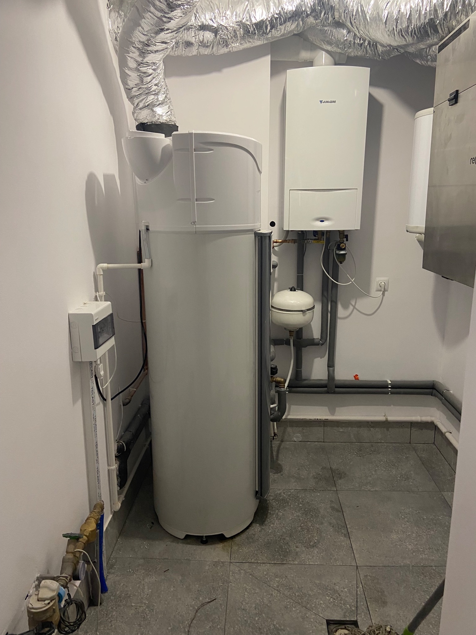 Biała instalacja grzewcza z zbiornikiem buforowym, rurami i kotłem Junkers na tle szarych płytek podłogowych i aluminiowej rury wentylacyjnej pod sufitem.
