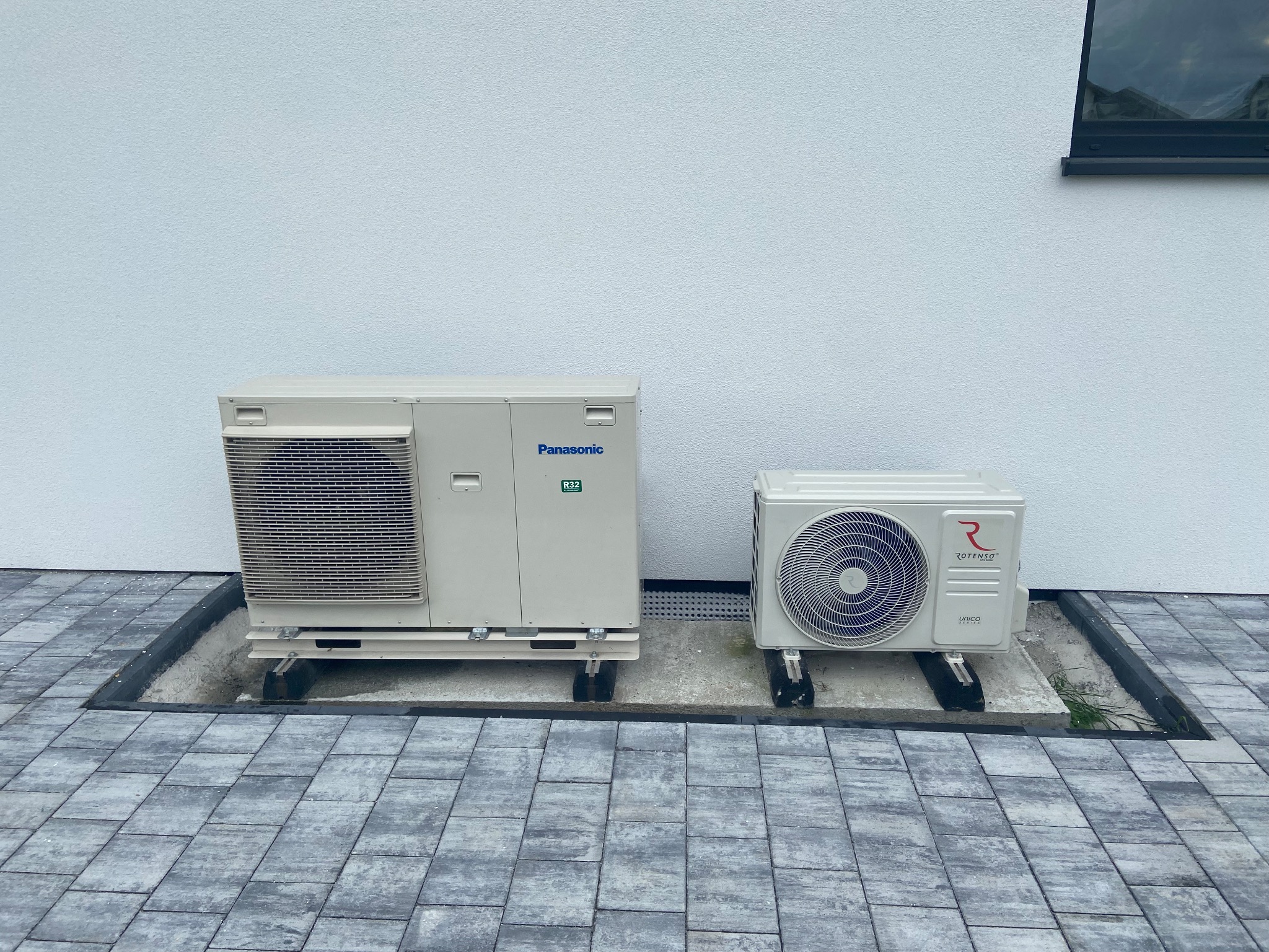 Zewnętrzne jednostki klimatyzacji Panasonic i Rotenso umieszczone na betonowych podstawach przy białej ścianie budynku, widoczne szare kostki brukowe.