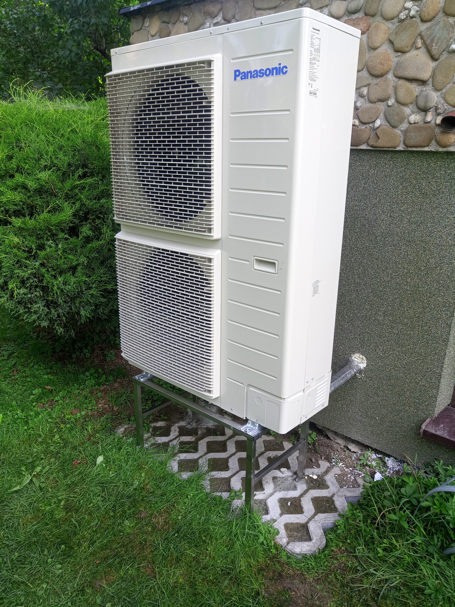 Zewnętrzna jednostka klimatyzatora Panasonic zamontowana na metalowym stelażu przy ścianie budynku z kamienną fakturą, otoczona zielenią.