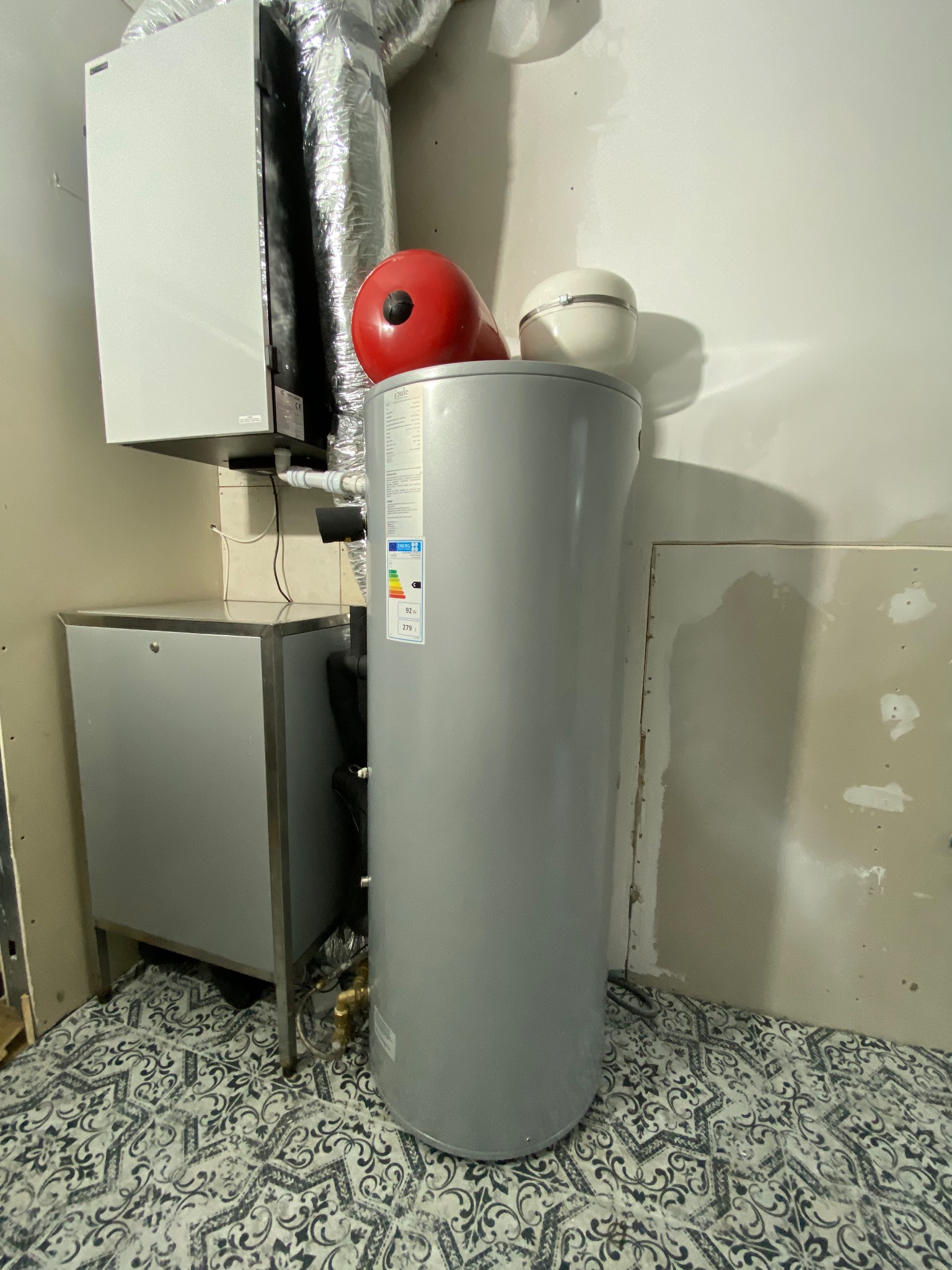 Szara, cylindryczna obudowa urządzenia z etykietą energetyczną, stojąca na wzorzystej podłodze w pomieszczeniu z elementami instalacji i niedokończonymi ścianami.