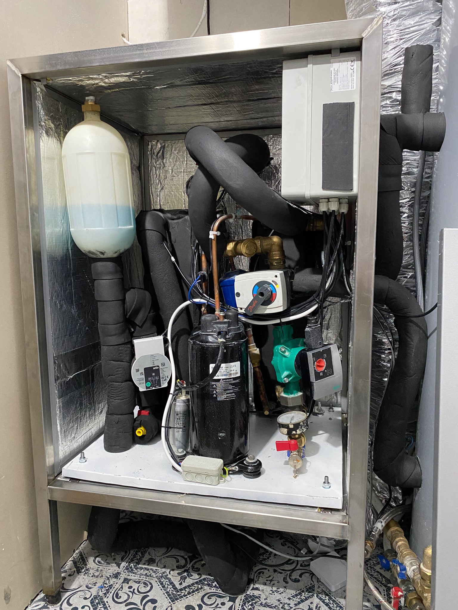 Kompletna instalacja w obudowie ze stali nierdzewnej, zawierająca zbiornik wyrównawczy, pompy obiegowe, zawory, przewody izolowane termicznie i sterowniki, umieszczona na wzorzystej podłodze.