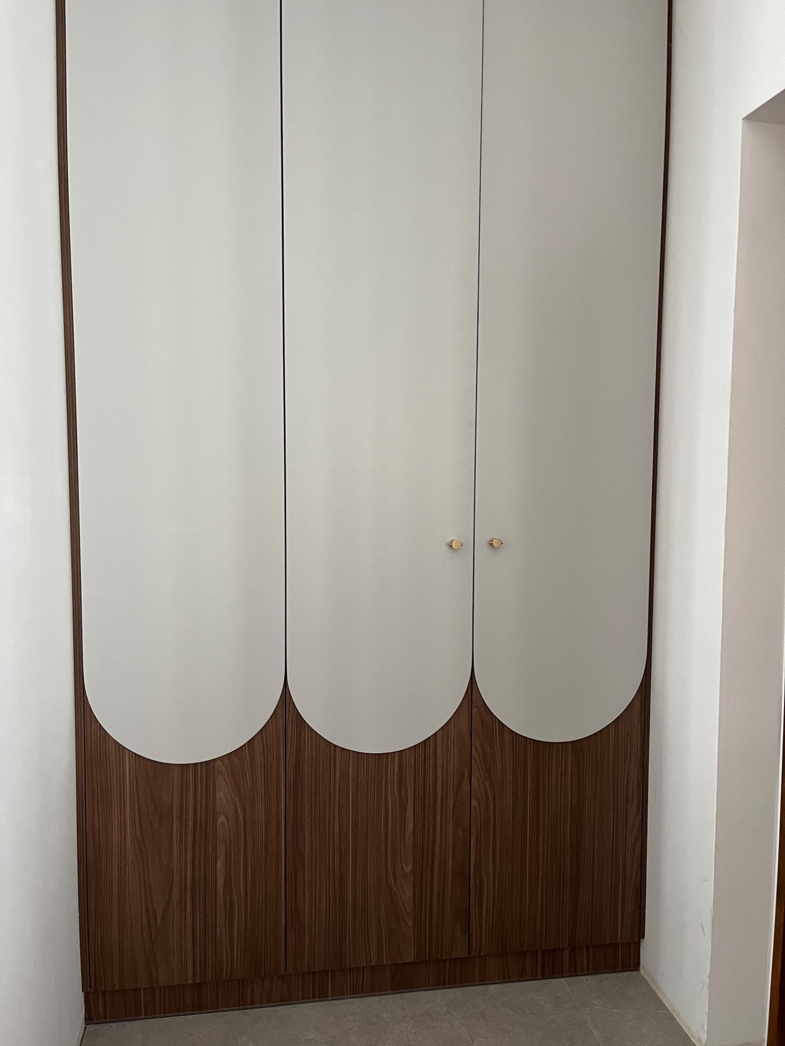 Trzydrzwiowa szafa z białymi frontami i dekoracyjnym, półokrągłym wykończeniem z forniru w kolorze ciemnego orzecha, z drobnymi, okrągłymi uchwytami.