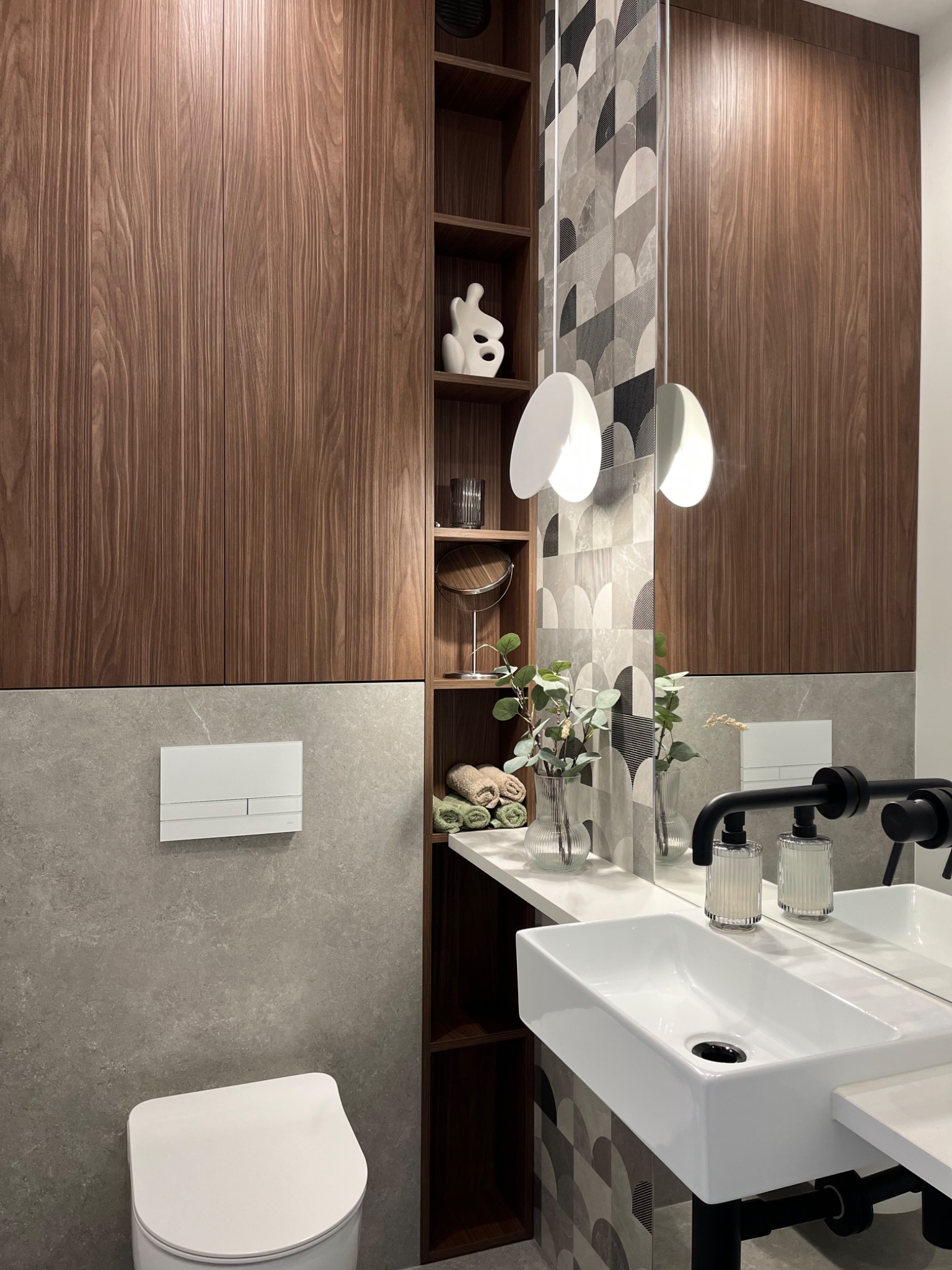 Nowoczesna łazienka z drewnianą zabudową, białą umywalką nablatową, armaturą w kolorze czarnym i dekoracyjnymi płytkami na ścianie.
