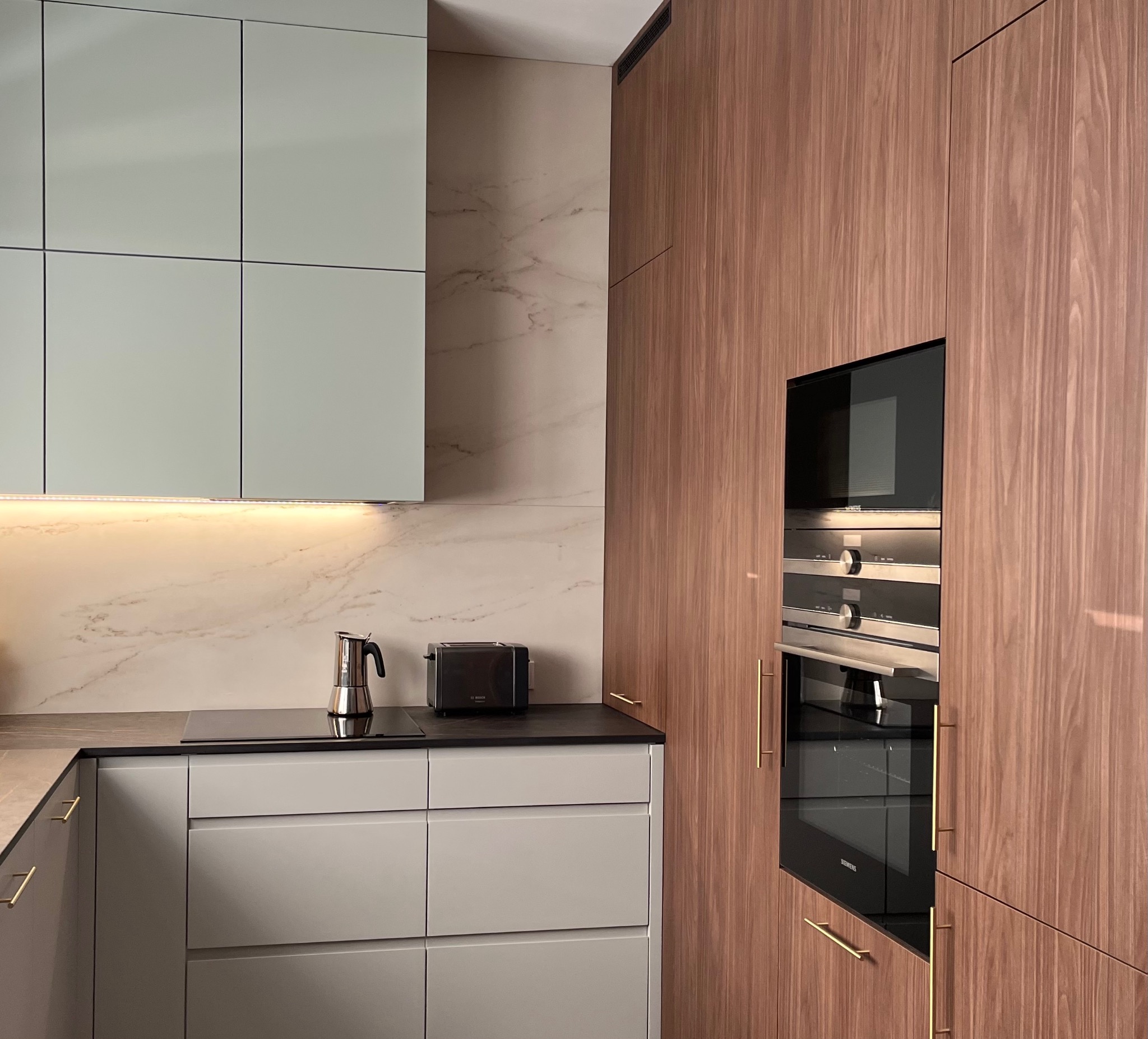 Nowoczesna kuchnia z szarymi szafkami i drewnianą zabudową piekarnika, uzupełniona o marmurowy backsplash i złote uchwyty.