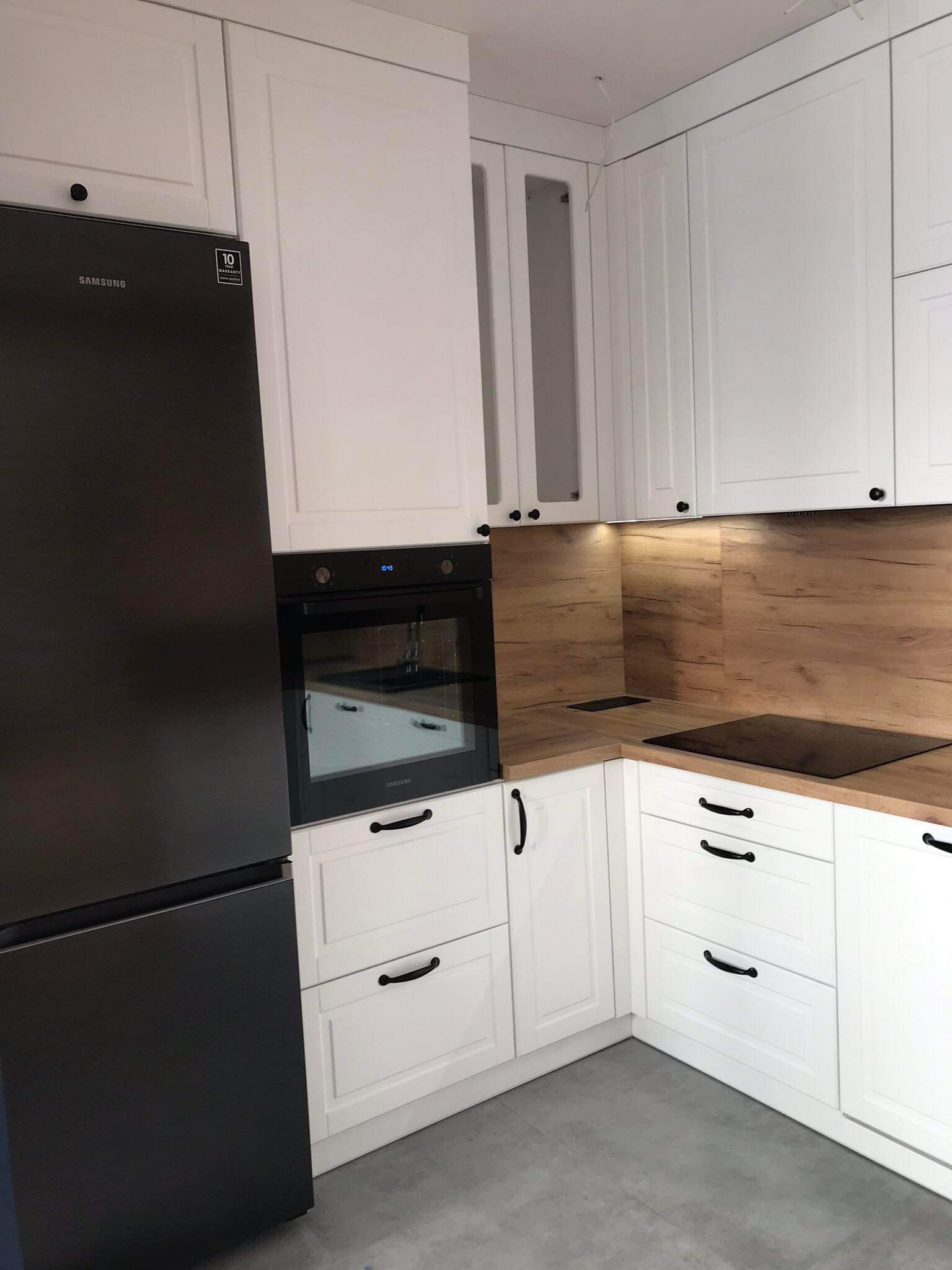 Nowoczesna kuchnia z białymi szafkami z frezowanymi frontami, drewnianym blatem i czarnym sprzętem AGD, w tym piekarnikiem i lodówką Samsung.