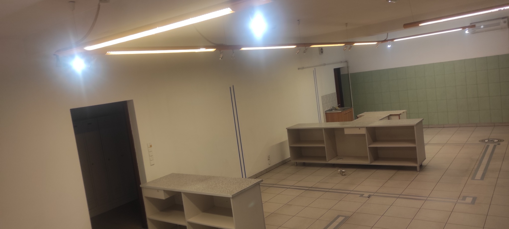 Wnętrze pustego lokalu biurowego z surowymi ścianami, podwieszonymi lampami LED i częściowo wyposażonymi stanowiskami pracy z białymi regałami.