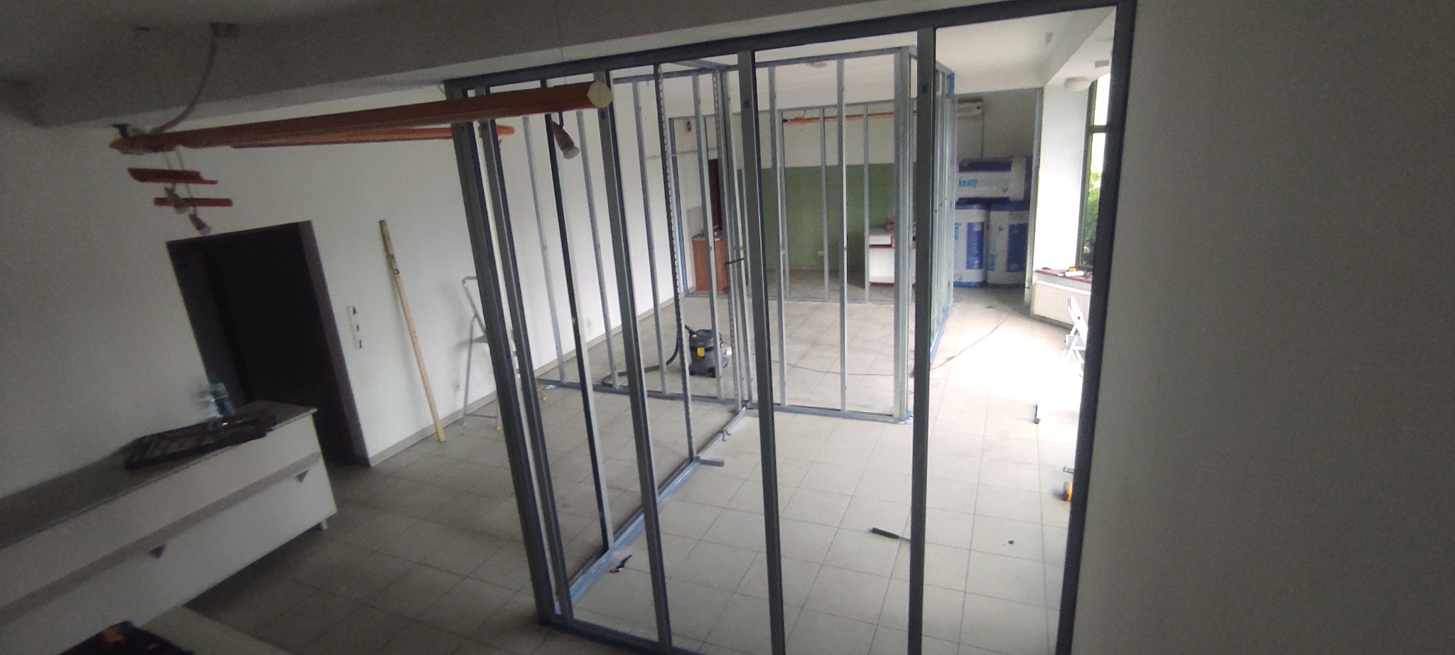 Stalowa konstrukcja szkieletowa ścian działowych w trakcie montażu w jasnym pomieszczeniu biurowym, widoczny odkurzacz przemysłowy i płyty izolacyjne w tle.