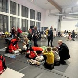 Er-Med Spółka z ograniczona odpowiedzialnością - Grupa osób ćwiczy resuscytację krążeniowo-oddechową na manekinach w przestronnej sali z dużymi oknami. Instruktor w czerwonym kombinezonie demonstruje technikę, a uczestnicy powtarzają czynności.