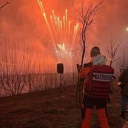 Er-Med Spółka z ograniczona odpowiedzialnością - Ratownik medyczny w pomarańczowym kombinezonie z napisem 'RATOWNIK' na plecach, obserwujący pokaz sztucznych ogni w tle, z widocznym nagłośnieniem i drzewami bez liści.