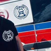 Fragment pojazdu z logo firmy ER-MED NZOZ, widoczne także na koszulce osoby stojącej obok. Ambulans z czerwono-biało-niebieskimi pasami.