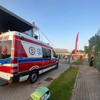 Ambulans z napisem 'AMBULANS' i symbolem róży w tle, zaparkowany na zewnątrz, prawdopodobnie podczas wydarzenia plenerowego, z widoczną sceną i publicznością w oddali.