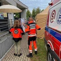 Ratownik medyczny w pomarańczowym uniformie, stojący tyłem do kamery obok ambulansu z widocznym symbolem ratownictwa medycznego, w tle budynek i rozłożony namiot.