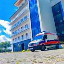 Ambulans zaparkowany przed nowoczesnym budynkiem w Swadzimiu, prawdopodobnie szpitalem lub kliniką. Widoczny fragment trawnika i chodnika. Karetka ma niebiesko-czerwone oznaczenia.