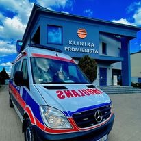 Ambulans z napisem 'Ambulans' odwróconym lustrzanie, zaparkowany przed budynkiem 'Klinika Promienista' z logo słońca, błękitne niebo z chmurami w tle.