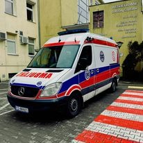 Ambulans Mercedes-Benz Sprinter z napisem 'REANIMACJA' zaparkowany przed budynkiem szpitala, widoczne detale karoserii i oznaczenia służby zdrowia.